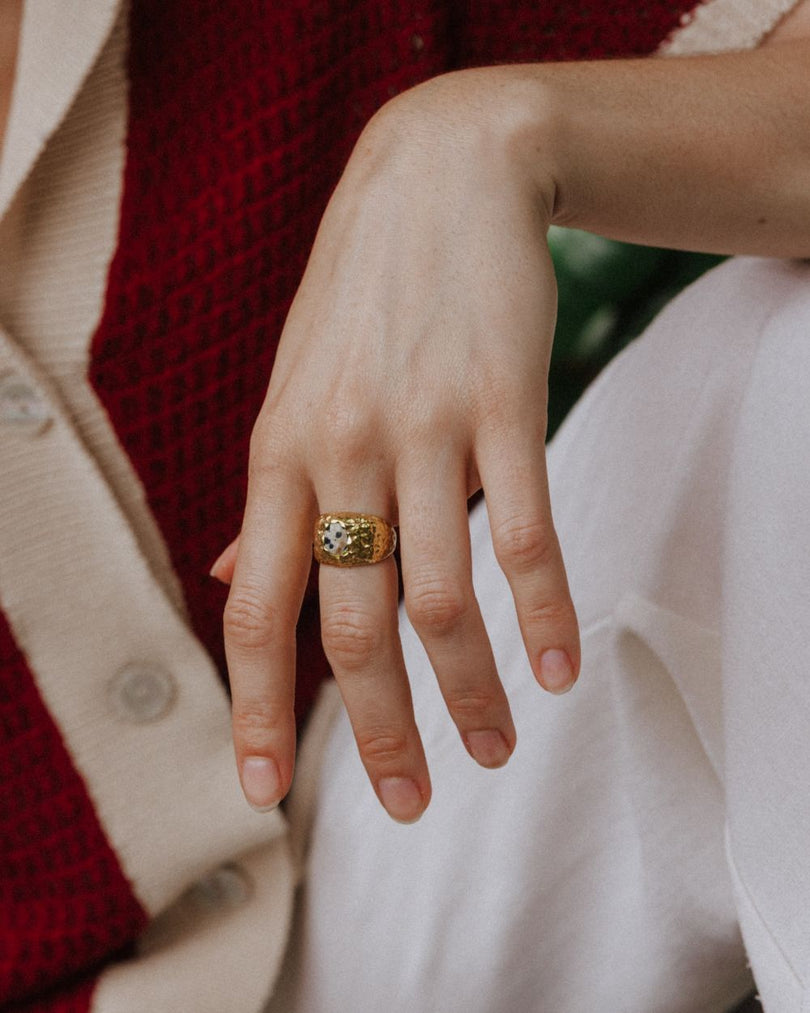 Primer plano de la modelo sentada mostrando la mano con el anillo bañado en oro de 18 quilates con piedra semipreciosa Jaspe Dálmata de Isla Vurma