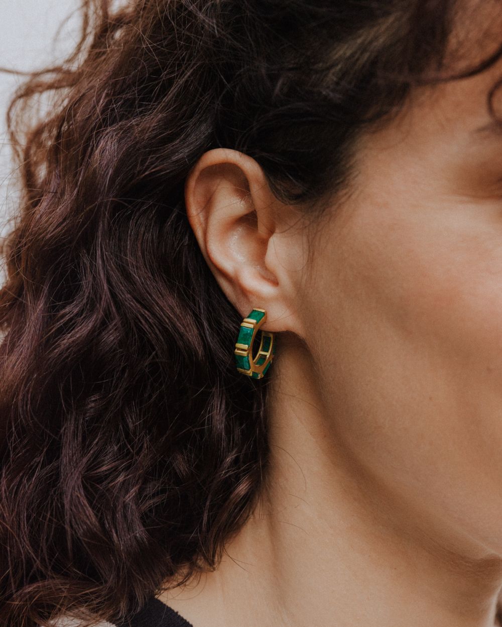Pendiente de aro dorado con piedras semipreciosas rectangulares en Esmeralda, lucido en la oreja de la modelo de cabello oscuro y rizado.