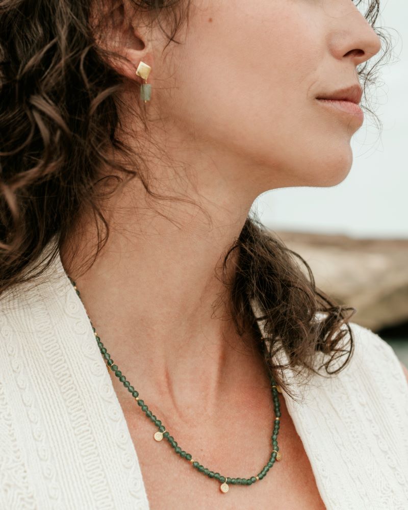 Mujer con cabello ondulado lleva un collar de cuentas Jade con pequeños colgantes dorados y pendientes geométricos dorados con piedra Aventurina Verde de Isla Vurma