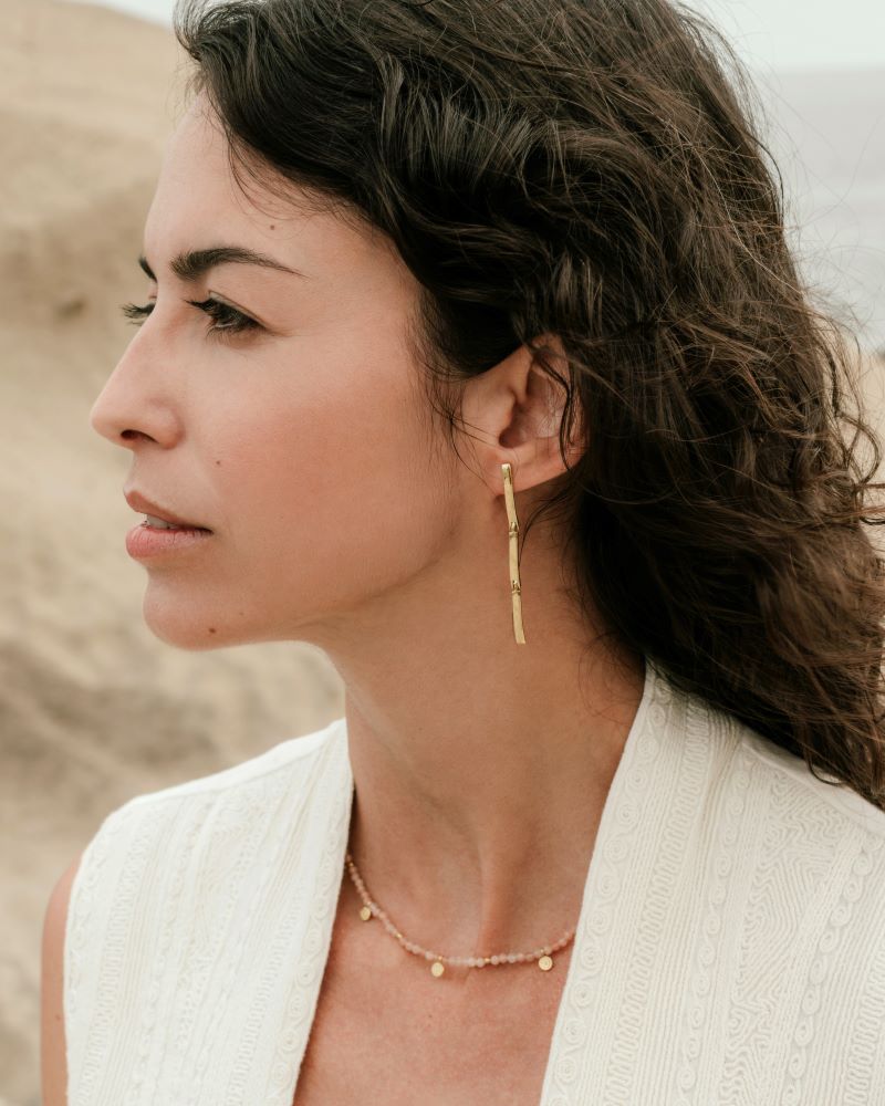 Mujer con el cabello oscuro y suelto lleva un collar de cuentas de piedras semipreciosas Piedra de Luna Melocotón con pequeños colgantes dorados y pendientes largos dorados de diseño minimalista de Isla Vurma