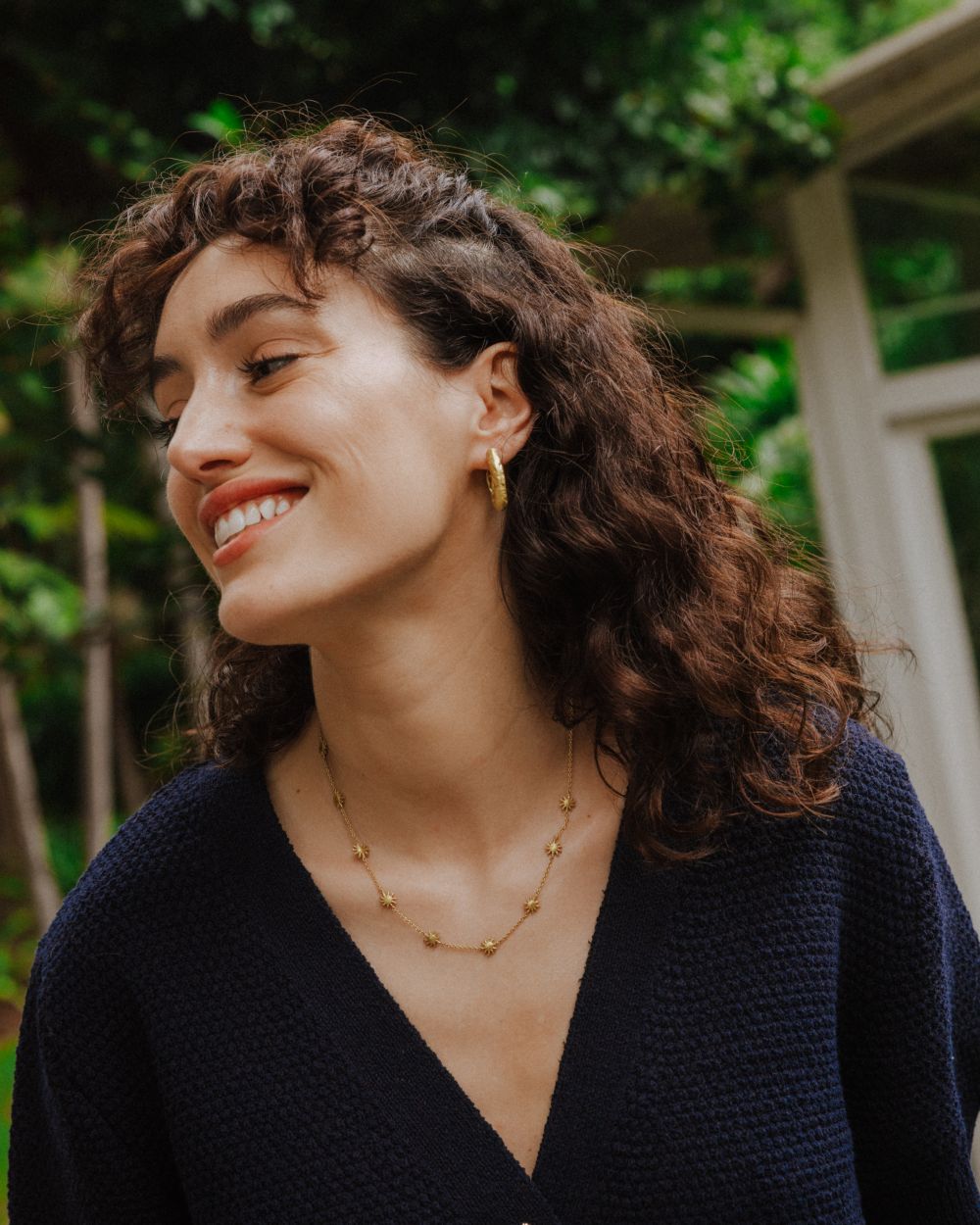 Retrato de la modelo sonriendo y mirando hacía un lado, que lleva un colla dorado con pequeñas estrellas distribuidas a lo largo de la cadena y unos aros dorados texturizados. Viste un jersey azul marino y el fondo muestra vegetación verde en un entorno natural.