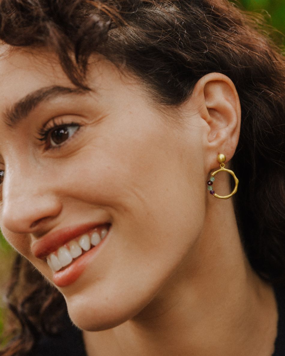Primer plano de una persona sonriendo, llevando un pendiente dorado con forma circular irregular y una pequeña sección de cuentas redondas en piedras semipreciosas Amatista, Aqua Calcedonia, Aventurina Verde y Rubí. Fondo natural desenfocado.
