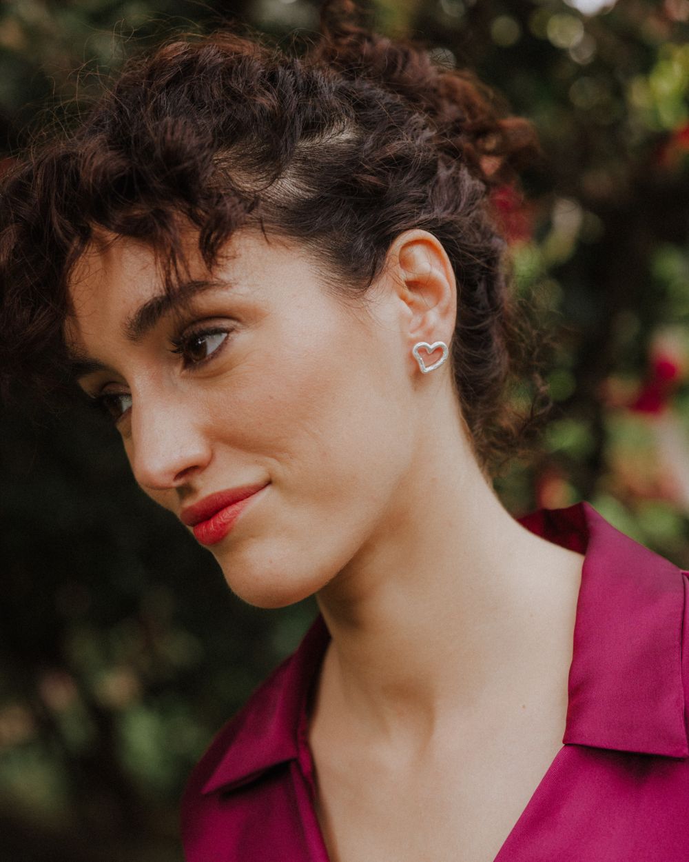 Primer plano de una persona con la cabeza ligeramente inclinada, llevando un pendiente plateado con forma de corazón irregular y diseño abierto ajustado al lóbulo. El fondo muestra vegetación y flores desenfocadas.