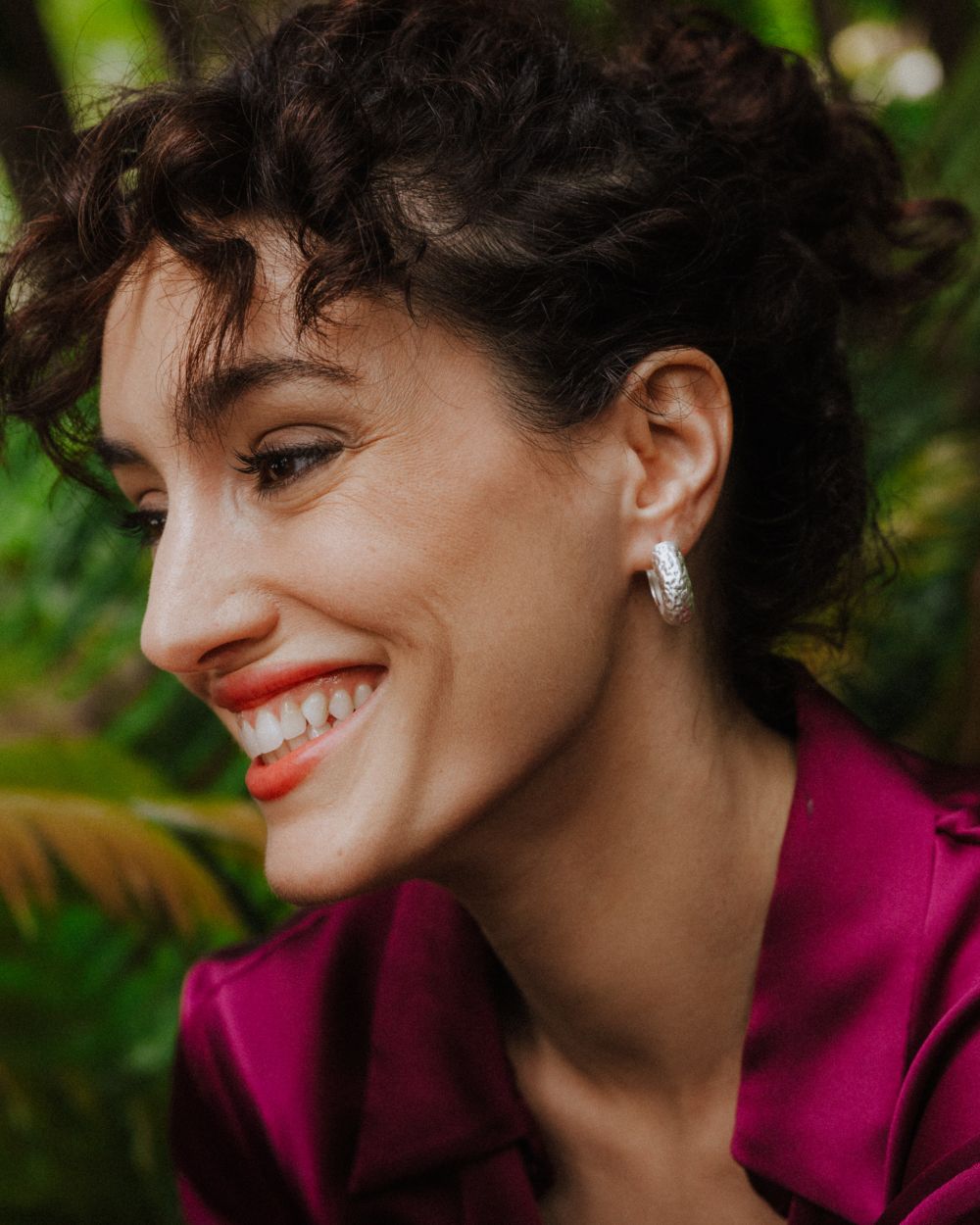 Primer plano de la modelo sonriente luciendo aros plateados de textura martillada. Lleva una blusa color magenta y se encuentra en un entorno natural con vegetación verde desenfocada al fondo.
