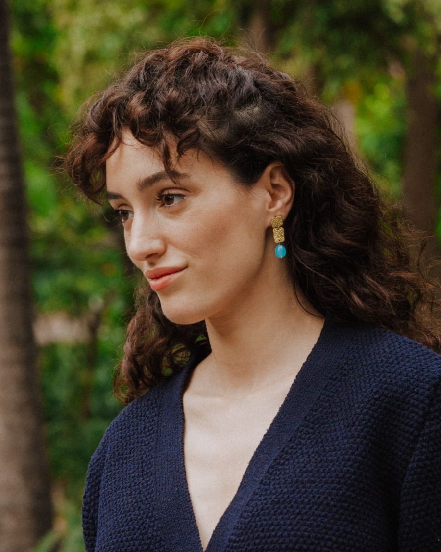 Pendientes dorados con dos piezas rectangulares texturizadas y una cuenta ovalada Aqua Calcedonia colgante, lucidos por una modelo en un entorno natural con vegetación de fondo.