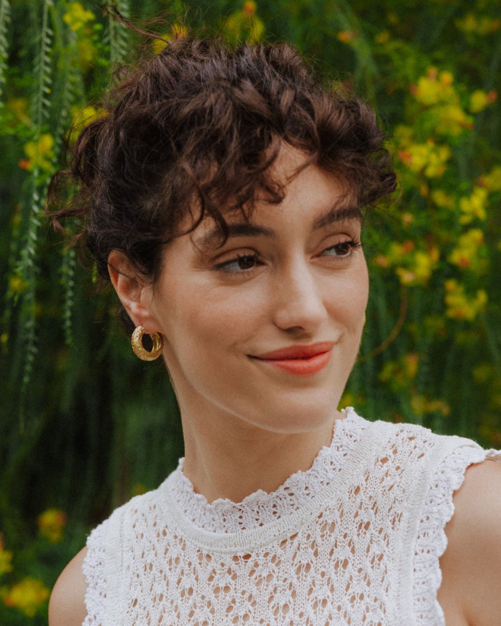 Pendientes dorados de aro grueso con textura martillada lucidos por una modelo, que lleva un top blanco de encaje. El fondo muestra un entorno natural con vegetación verde y flores amarilla.