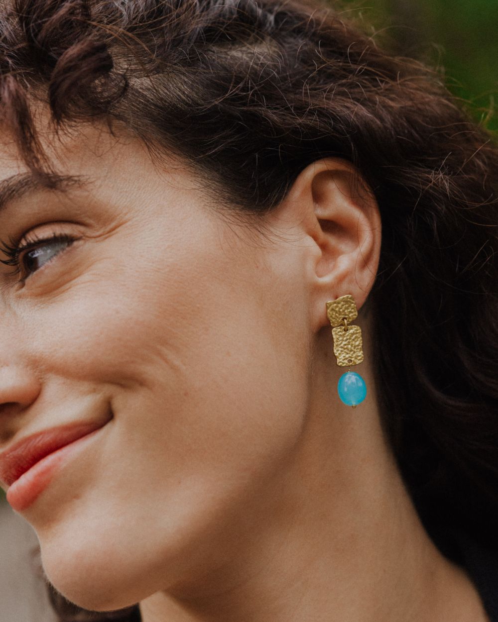 Pendiente dorado colgante formado por dos piezas rectangulares texturizadas y una cuenta en piedra semipreciosa Aqua Calcedonia al final. La pieza se aprecia en la oreja de una modelo, con un fondo natural y tonos verdes desenfocados.