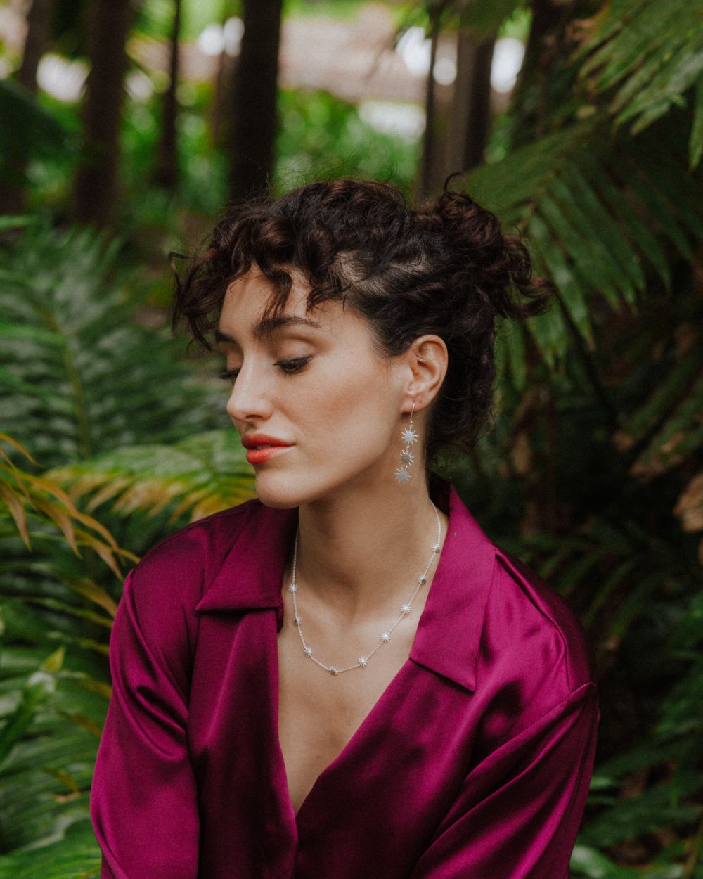 Persona sentada en un entorno natural con vegetación, llevando pendientes largos plateados formados por tres pequeñas piezas en forma de sol de bordes irregulares y rayos finos, unidas entre sí con pequeñas argollas. Los pendientes cuelgan desde un gancho fino. La persona viste una camisa satinada color magenta y también lleva un collar plateado con pequeñas piezas en forma de sol. Fondo de plantas desenfocado.