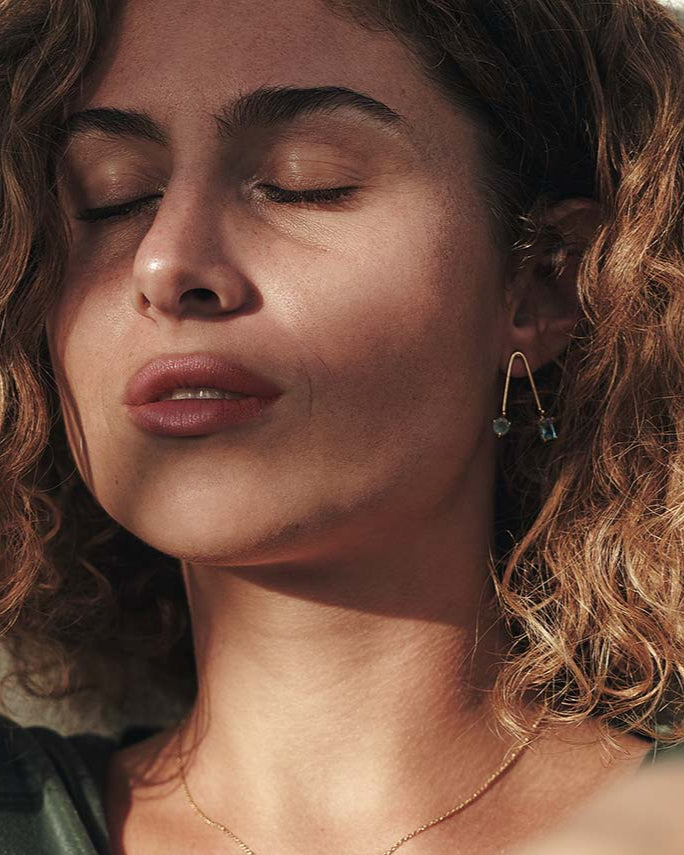 Mujer con pendientes de latón bañados en oro 18k artesanales de latón dorado con piedras apatite y aqua calcedonia. Diseño elegante y minimalista, perfecto para ocasiones especiales o para un look sofisticado diario.