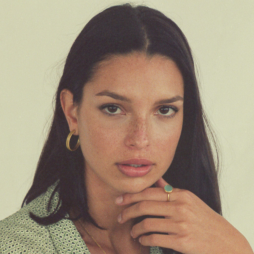 Pendientes de aro dorados en oreja de modelo con cabello largo y camisa verde claro, fondo neutro