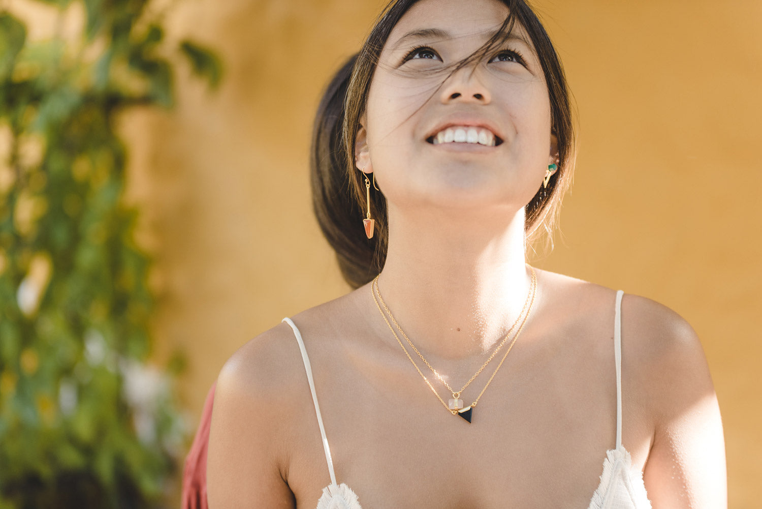 Modelo con conjunto de collares bañados en oro de 18 quilates con piedra semipreciosa de Cuarzo Rosa sujeta por una pequeña pieza dorada y una pequeña pieza triangular negra, además de unos pendientes en forma triangular dorados y otros con la piedra semipreciosa Jade de Isla Vurma