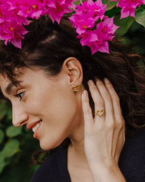 Primer plano de una persona de perfil sonriendo, llevando un anillo dorado con diseño abierto en forma de corazón irregular y un pendiente a juego en la oreja. La mano está apoyada cerca del rostro y, sobre la cabeza, se ven flores rosas en primer plano con hojas verdes desenfocadas al fondo.