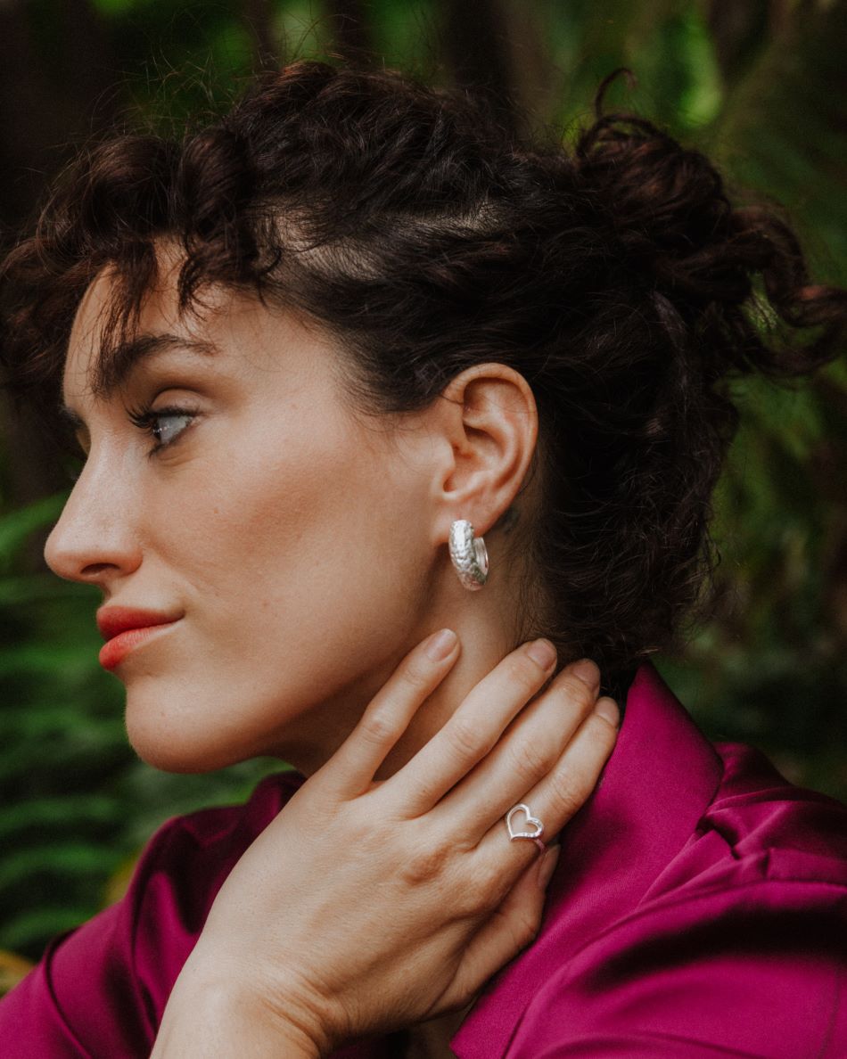 Primer plano de una persona de perfil con la mano apoyada en el cuello, llevando un anillo plateado de diseño abierto en forma de corazón irregular. También se aprecia un pendiente plateado de aro grueso en la oreja. Fondo natural desenfocado con tonos verdes.