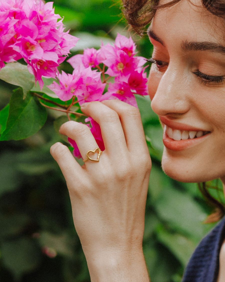 Primer plano de una mano acercando flores rosas al rostro, mostrando un anillo dorado con diseño abierto en forma de corazón irregular. Se ve parte del rostro sonriendo y un fondo de hojas verdes y flores desenfocadas.