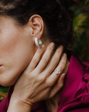 Primer plano de una mano apoyada en el cuello mostrando un anillo plateado con diseño abierto en forma de corazón irregular. También se ve un pendiente plateado de aro grueso en la oreja. Fondo natural desenfocado con tonos verdes.