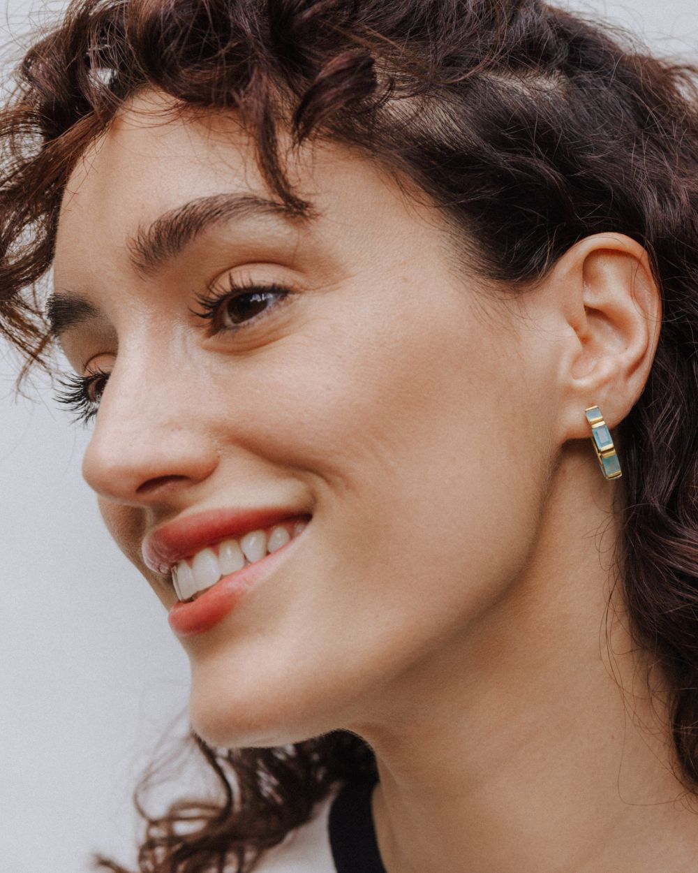 Primer plano de la modelo sonriendo que lleva un pendiente de aro dorado con piedras semipreciosas rectangulares en aqua calcedonia. En fondo claro.