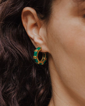 Primer plano de un pendiente de aro dorado con piedras semipreciosas rectangulares en Esmeralda, colocado en la oreja de una mujer de cabello oscuro.