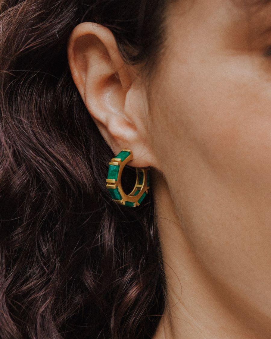 Primer plano de un pendiente de aro dorado con piedras semipreciosas rectangulares en Esmeralda, colocado en la oreja de una mujer de cabello oscuro.