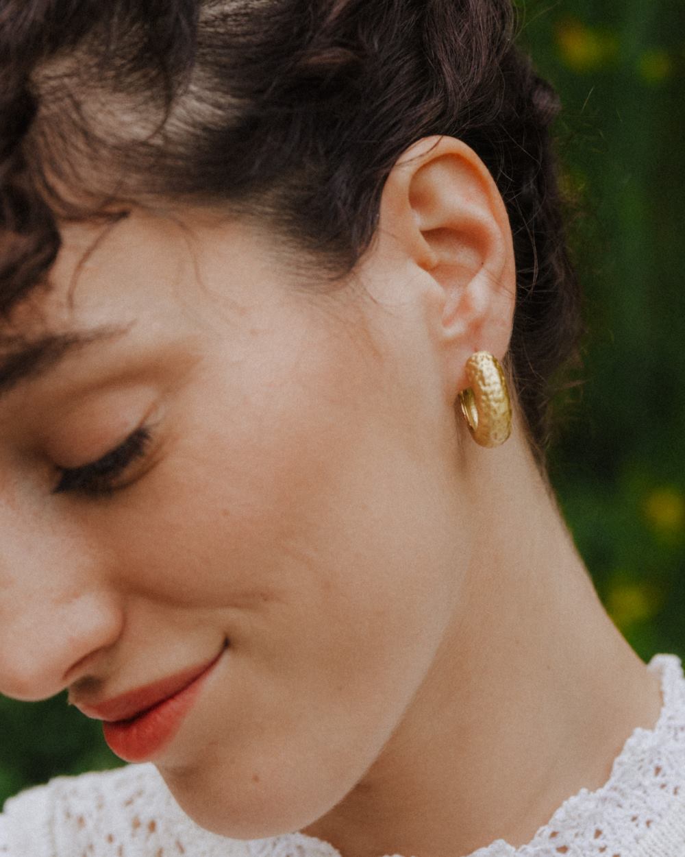 Primer plano de una modelo inclinando la cabeza hacia abajo, luciendo aros dorados de textura martillada. Se aprecia su top blanco de encaje y un fondo verde desenfocado con tonos amarillos.