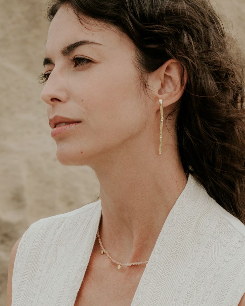 Mujer de cabello oscuro y ondulado lleva un collar de cuentas Piedra de Luna Melocotón con pequeños colgantes dorados y unos pendientes largos geométricos dorados de Isla Vurma