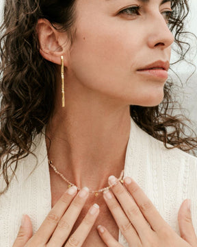 Mujer con cabello rizado sostiene con sus manos un collar de cuentas con piedras semipreciosas de Piedra de Luna Melocotón con pequeños colgantes dorados y luce pendientes largos dorados de Isla Vurma
