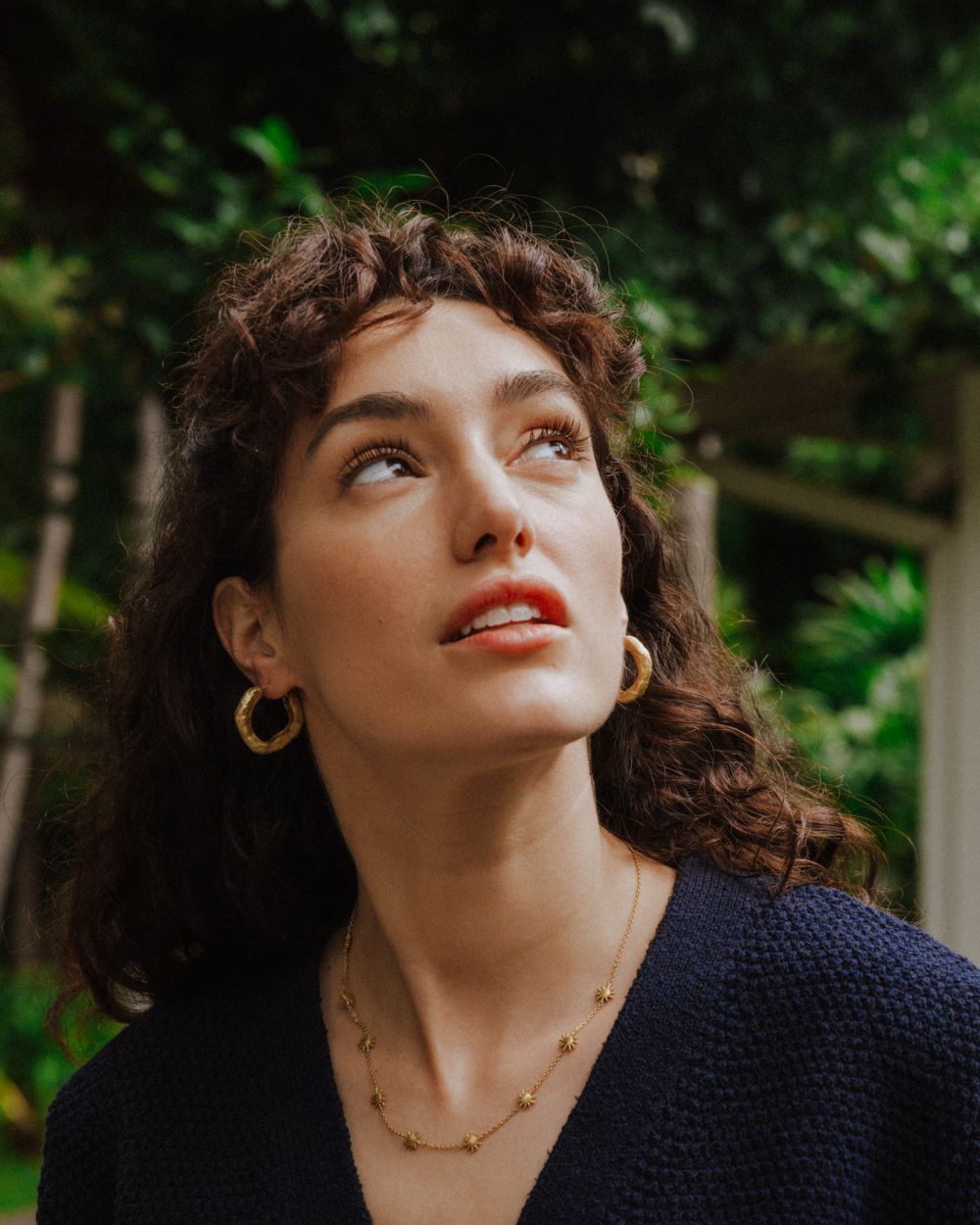 Retrato de la modelo con el rostro inclinado hacía arriba, luciendo un collar dorado con pequeños colgantes en forma de estrella y unos aros gruesos dorados de textura martillada