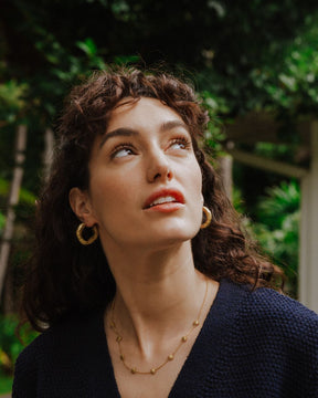 Retrato de la modelo con el rostro inclinado hacía arriba, luciendo un collar dorado con pequeños colgantes en forma de estrella y unos aros gruesos dorados de textura martillada