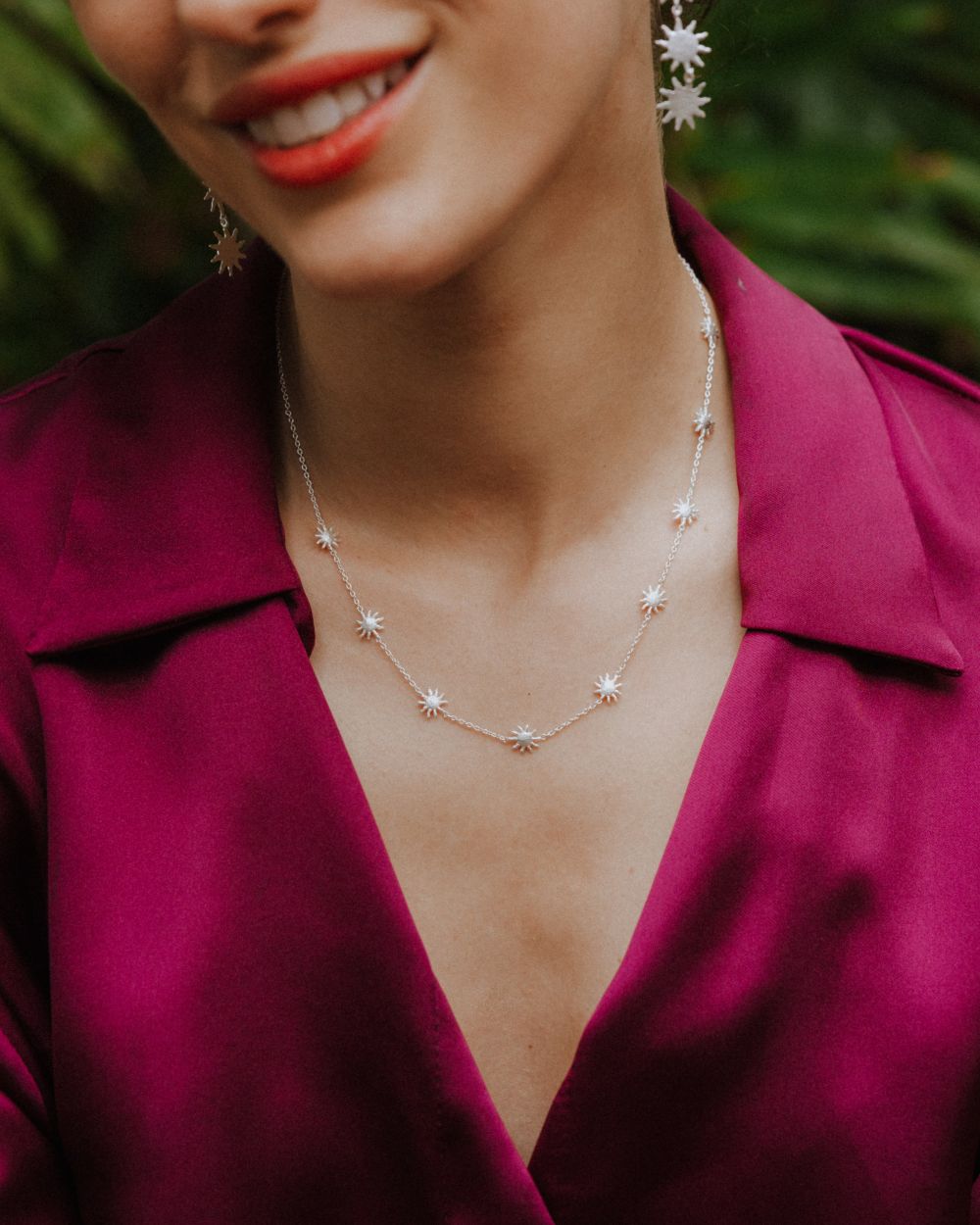 Primer plano de la modelo sonriendo que lleva un collar plateado con pequeñas estrellas distribuidas a lo largo de una cadena fina. Viste una blusa satinada de color fucsia y pendientes a juego con forma de estrella.