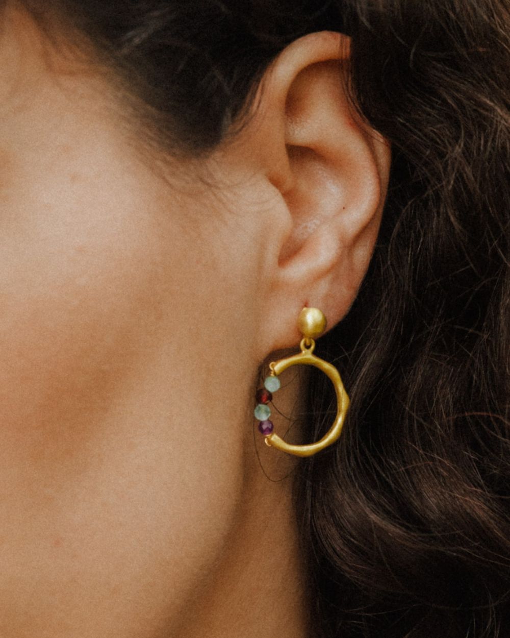 Primer plano de una oreja llevando un pendiente dorado con forma circular irregular y una pequeña sección de cuentas redondas en piedras semipreciosas Amatista, Aqua Calcedonia, Aventurina Verde y Rubí. El pendiente tiene cierre de tuerca.