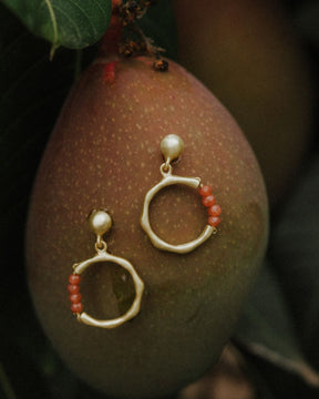 Pendientes dorados con forma circular irregular y bolita superior como cierre. En la parte inferior de cada aro aparece una fila de pequeñas cuentas redondas en piedras semipreciosas Cuarzo Meloctón. Están apoyados sobre la superficie de un fruto verde-rojizo, rodeados de hojas naturales en un ambiente vegetal.