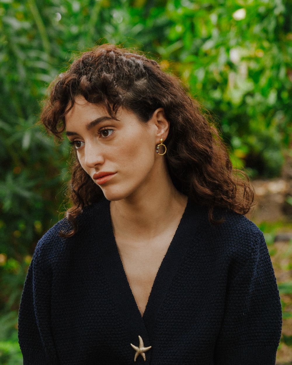 Primer plano de una mujer con pendientes dorados en forma de aro irregular con pequeñas cuentas de piedras semipreciosas en un entorno natural. Lleva un jersey azul marino con un broche en forma de estrella de mar, y el fondo muestra vegetación verde.