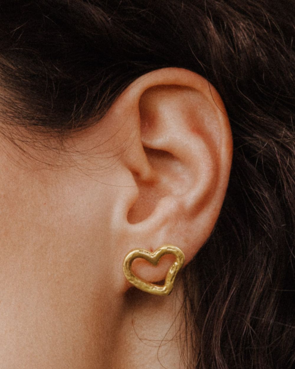 Primer plano de una oreja llevando un pendiente dorado con forma de corazón irregular y diseño abierto. El pendiente queda ajustado al lóbulo.