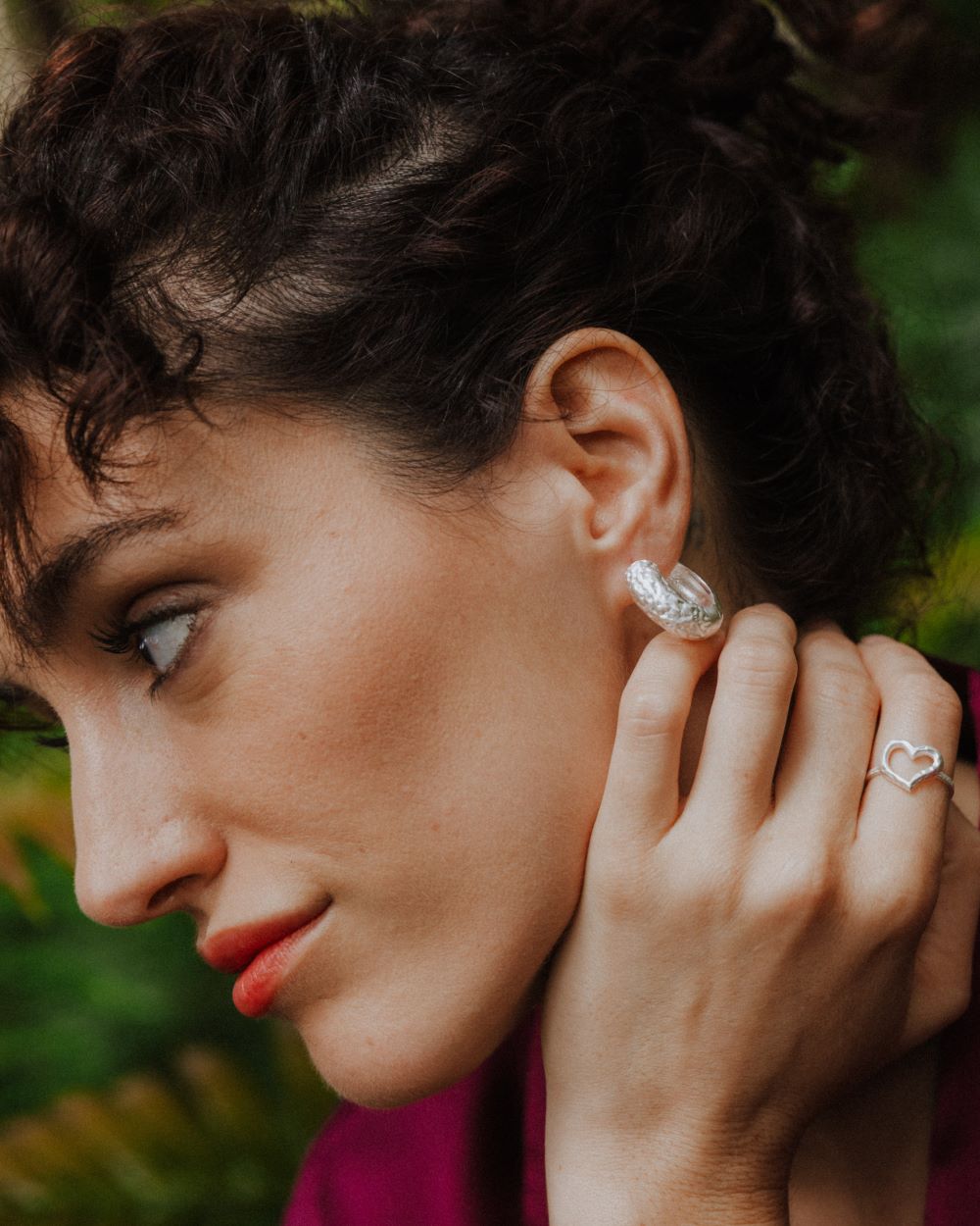 Primer plano de una mujer con labios rojos y blusa color magenta, luciendo aros plateados de textura martillada y un anillo en forma de corazón. Fondo natural verde desenfocado.