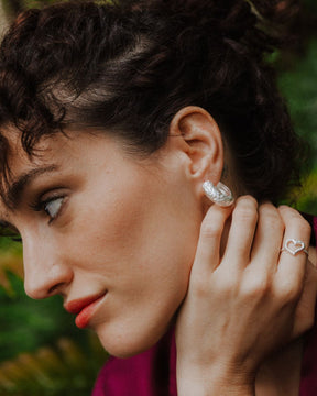Primer plano de una mujer con labios rojos y blusa color magenta, luciendo aros plateados de textura martillada y un anillo en forma de corazón. Fondo natural verde desenfocado.