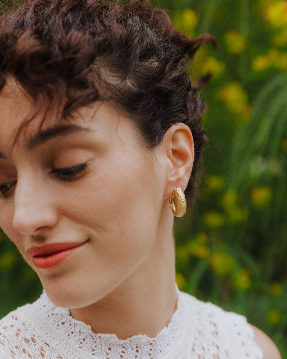 Primer plano de una modelo luciendo unos aros dorados de grosor medio con textura martillada. Lleva un top blanco de encaje y el fondo muestra vegetación verde con pequeñas flores amarillas.