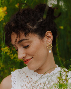 Pendientes dorados asimétricos con perla Natural, lucidos por la modelo al aire libre con vegetación de fondo
