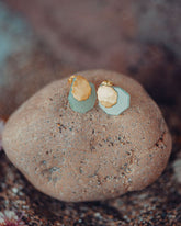 Pendientes pequeños de latón bañado en oro 18k con una piedra tallada en Aventurina Verde en forma geométrica, colocados sobre una roca de tonos marrones con pequeñas motas oscuras.