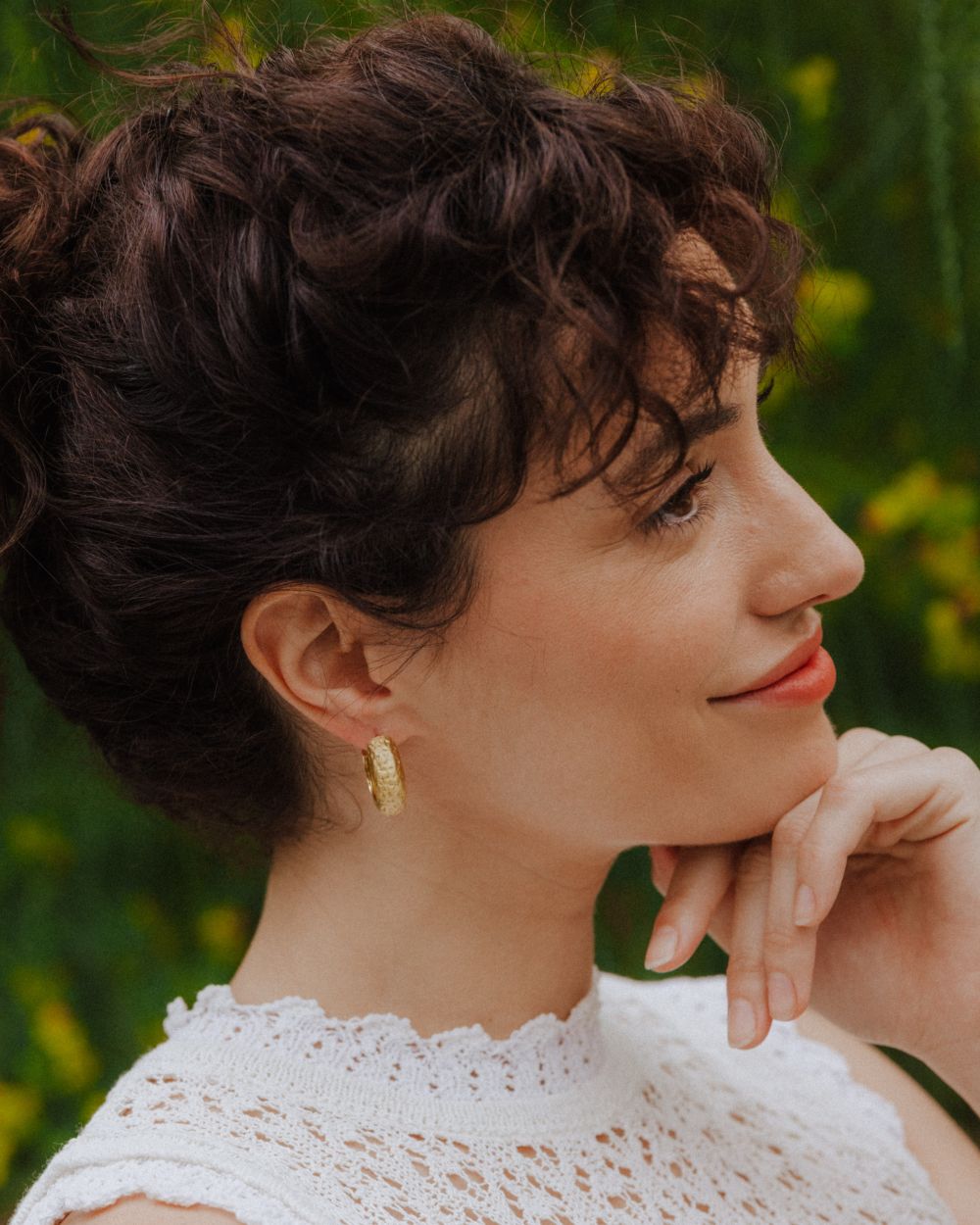 Pendiente tipo aro dorado con textura orgánica colocado en la oreja, acompañado de un gesto relajado con la mano apoyada en el mentón. La persona viste un top blanco de encaje y el fondo verde natural.
