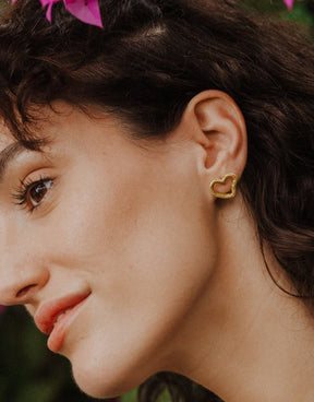 Primer plano de una persona de perfil inclinando ligeramente la cabeza, llevando un pendiente dorado con forma de corazón irregular y diseño abierto ajustado al lóbulo. Se aprecian flores rosas desenfocadas en la parte superior y un fondo vegetal en tonos verdes.