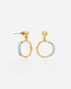 Primer plano de unos pendientes dorados con forma circular irregular y cierre de tuerca con bolita redonda. En la parte baja de cada aro cuelga una fila de pequeñas cuentas en piedra semipreciosa Aqua Calcedonia. Fondo liso y claro.