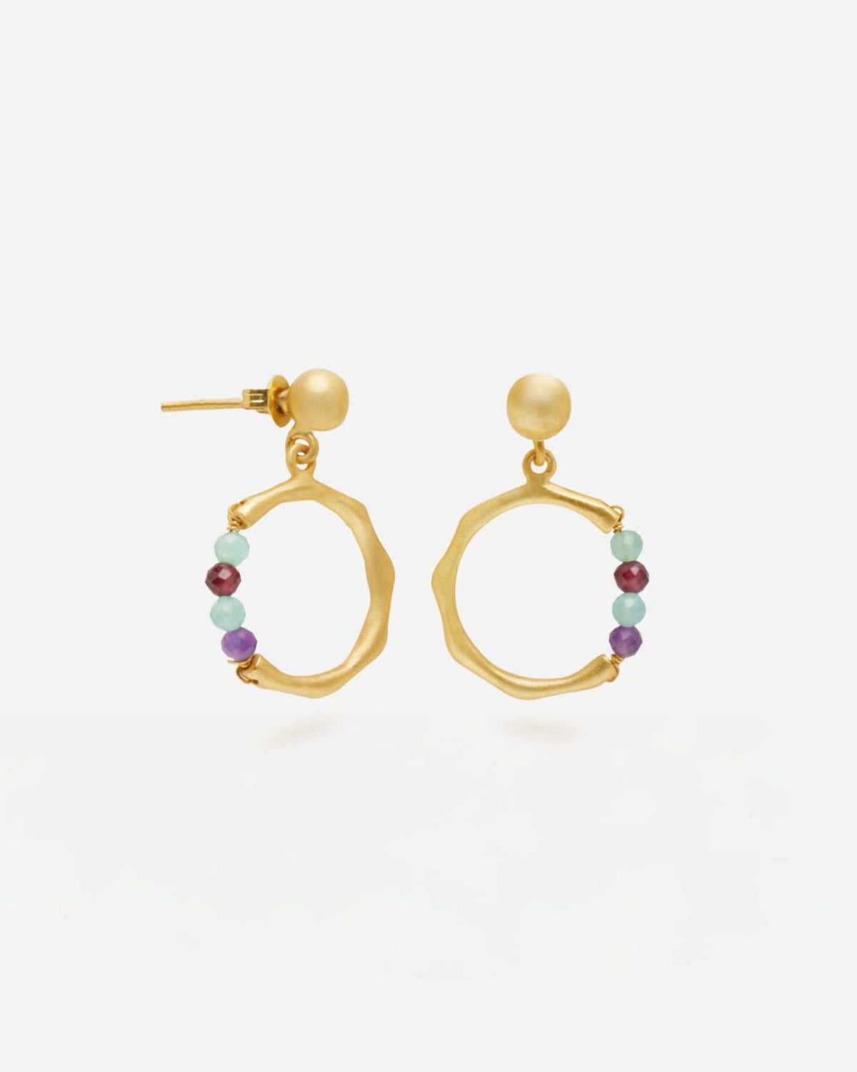 Primer plano de unos pendientes dorados con forma circular irregular y cierre de tuerca. En la parte inferior de cada aro cuelga una pequeña sección de cuentas redondas en piedras semipreciosas Amatista, Aqua Calcedonia, Aventurina Verde y Rubí. Fondo liso y claro.