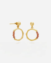 Primer plano de unos pendientes dorados con forma circular irregular y cierre de tuerca. En la parte inferior de cada aro cuelga una pequeña sección de cuentas redondas en piedras semipreciosas Cuarzo Melocotón. Fondo liso y claro.