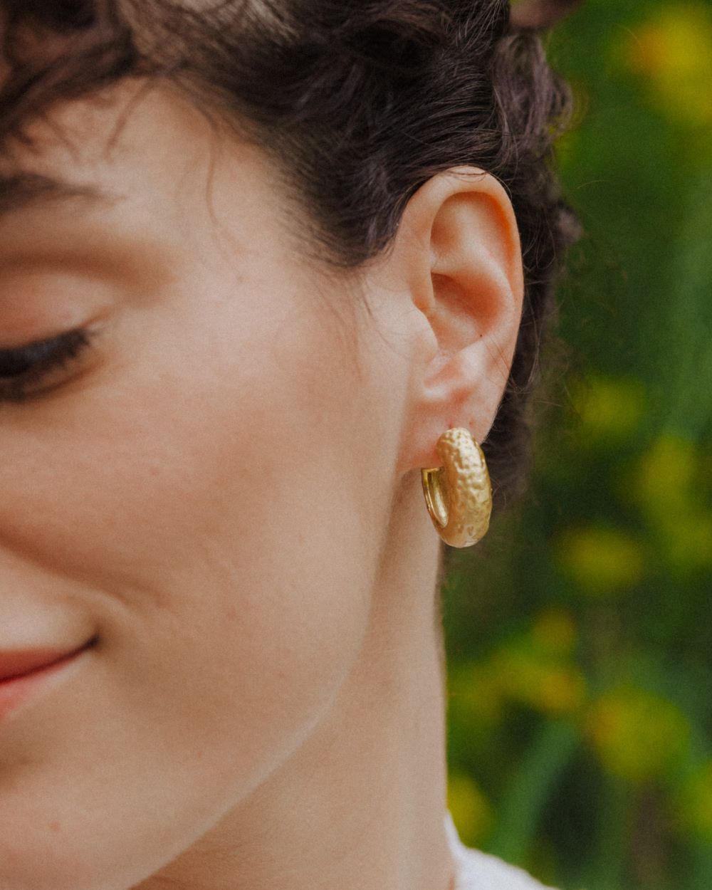 Primer plano de un aro dorado de grosor medio y textura martillada colocado en la oreja de una persona. Fondo natural y desenfocado en tonos verdes.