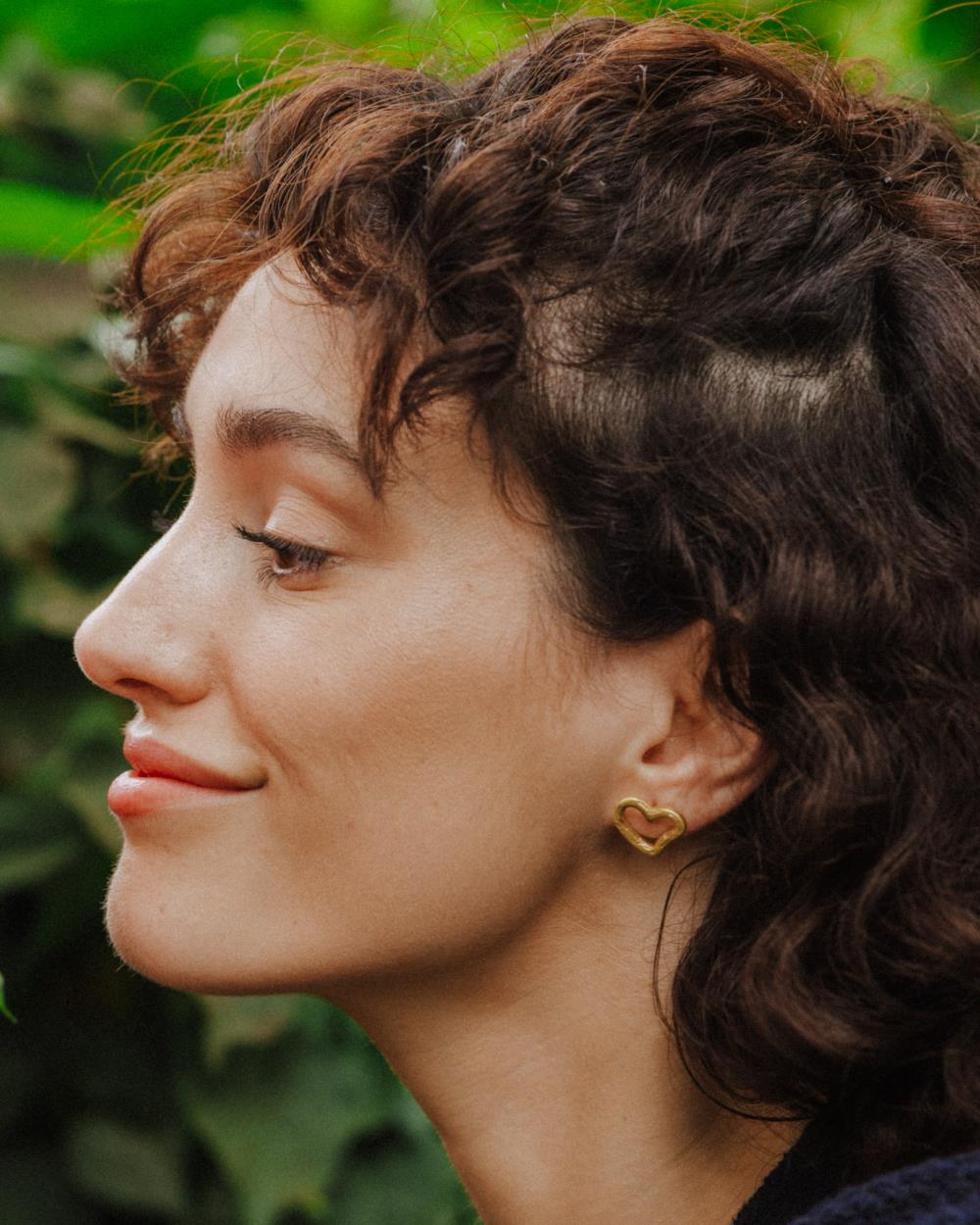 Primer plano de una persona de perfil sonriendo, llevando un pendiente dorado con forma de corazón irregular y diseño abierto ajustado al lóbulo. El fondo muestra hojas verdes desenfocadas.