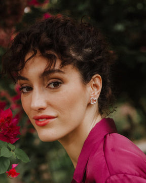 Primer plano de una persona mirando a cámara, llevando un pendiente plateado con forma de corazón irregular y diseño abierto ajustado al lóbulo. Se ven flores rosas desenfocadas en primer plano y un fondo natural en tonos verdes.