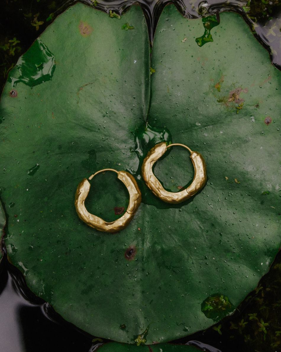 Primer plano de los pendientes gruesos circulares irregulares bañados en oro de 18k sobre una hoja verde y agua de Isla Vurma