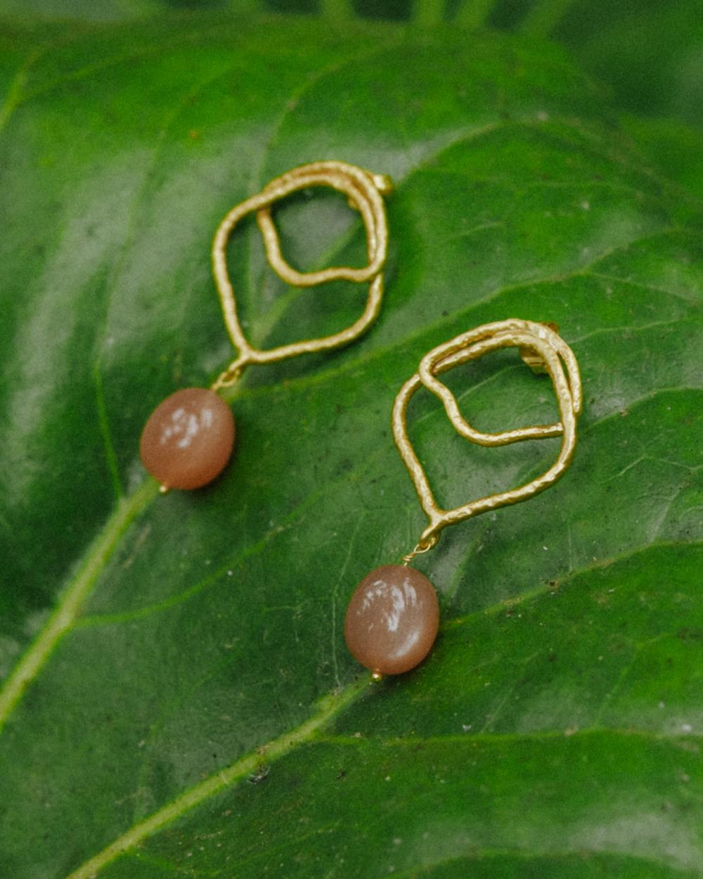 Primer planos de los pendientes asimétricos bañados en oro de 18 quilates con piedra semipreciosa Cuarzo Melocotón en forma de aceituna mini, Isla Vurma sobre hoja natural verde 