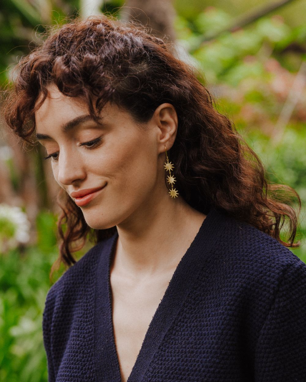 Primer plano de una persona con el pelo rizado mirando hacia abajo, llevando un pendiente largo dorado formado por tres pequeñas piezas en forma de sol de bordes irregulares y rayos finos, unidas entre sí con pequeñas argollas. El pendiente cuelga desde un gancho fino y destaca sobre el fondo natural desenfocado