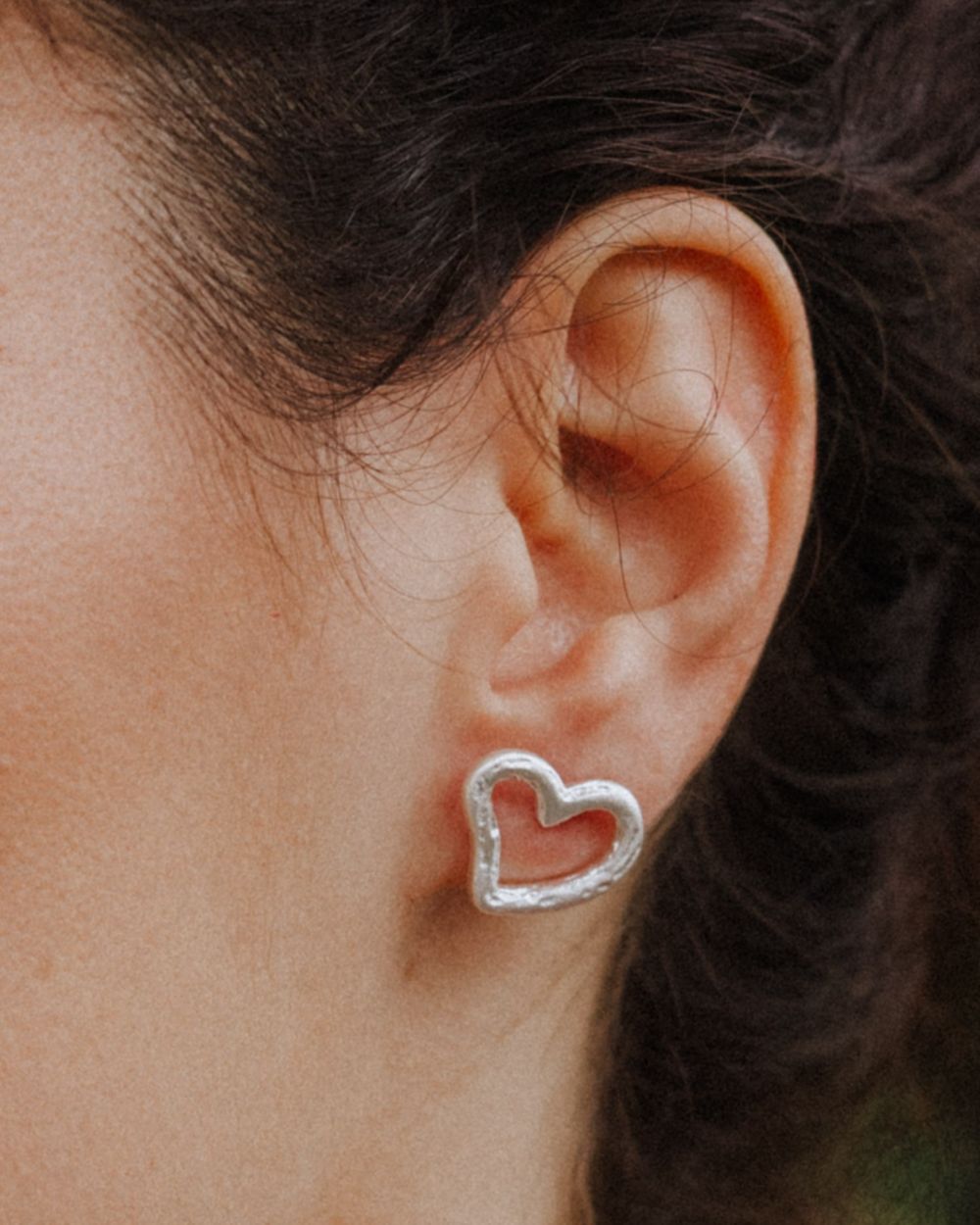 Primer plano de una oreja llevando un pendiente plateado con forma de corazón irregular y diseño abierto, ajustado al lóbulo. Fondo natural desenfocado.