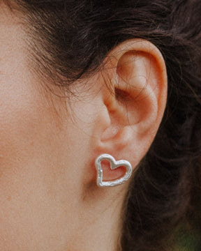 Primer plano de una oreja llevando un pendiente plateado con forma de corazón irregular y diseño abierto, ajustado al lóbulo. Fondo natural desenfocado.